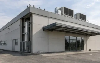 Angolo di un edificio commerciale visto dal piazzale, con il lungo cornicione del bordo del tetto, la tettoia metallica sopra l’ingresso vetrato e due unita di trattamento aria sulla copertura piana, con alcuni piccioni posati sul cornicione