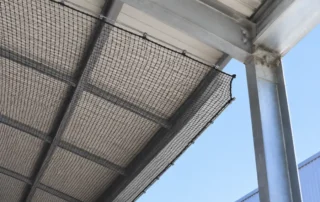 Rete anti-volatili tesa sotto la copertura metallica di una tettoia, ripresa dal basso, con un varco aperto lungo il bordo destro dove la rete non arriva alla trave