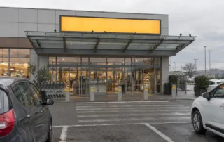 Ingresso di un supermercato con pensilina in vetro e acciaio e alcuni piccioni posati sul bordo superiore, sopra le porte automatiche