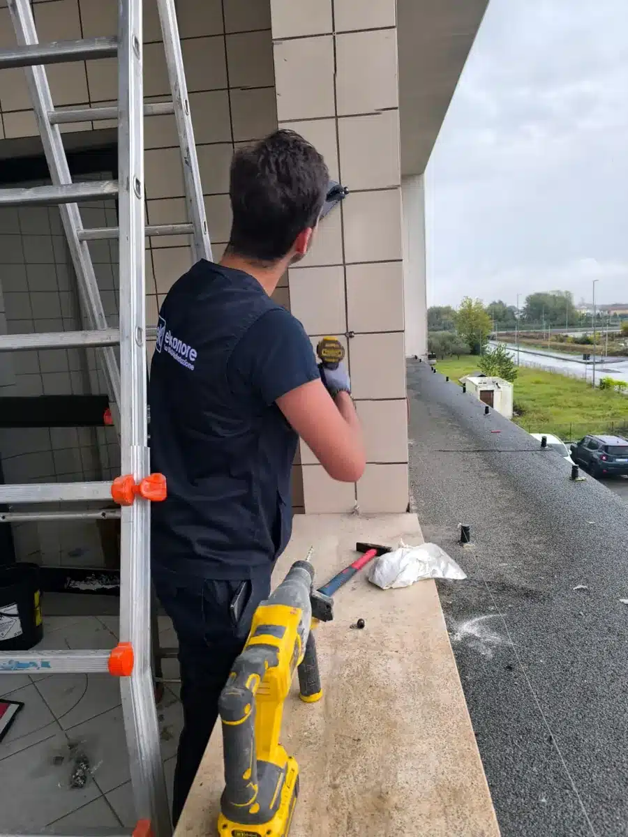 Tecnico Ekonore fissa i supporti dei dissuasori sulla facciata di un edificio, con scala e trapano